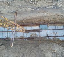 Ferraillage des semelles filantes. La ferraille touche le sol mais le maçon remarque que la cale n'est pas bien mise. Les ferrailles verticales sont fixées dans les côtés de la tranchée par un autre fer à béton.