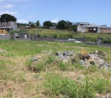 Vue depuis l'autre côté du terrain.
A gauche notre entrée, à droite celle de nos futurs voisins.
En face de l'autre coté de la voie ce sont également deux parcelles qui font partie de notre lotissement.
Il y a deux parcelles en dessous de la notre, donc très très peu de passage (le lotissement est une voie sans issue).
Nous allons être tranquilles :)
Sur la photo on voit bien les roches présentes sur le terrain (veine de roche dite "bleue"). C'est la roche la plus difficile à travailler ici à la Réunion, le budget terrassement sera plus important que pour un terrain "meuble", en revanche pas de mur de soutènement à prévoir, et ça c'est un gros .